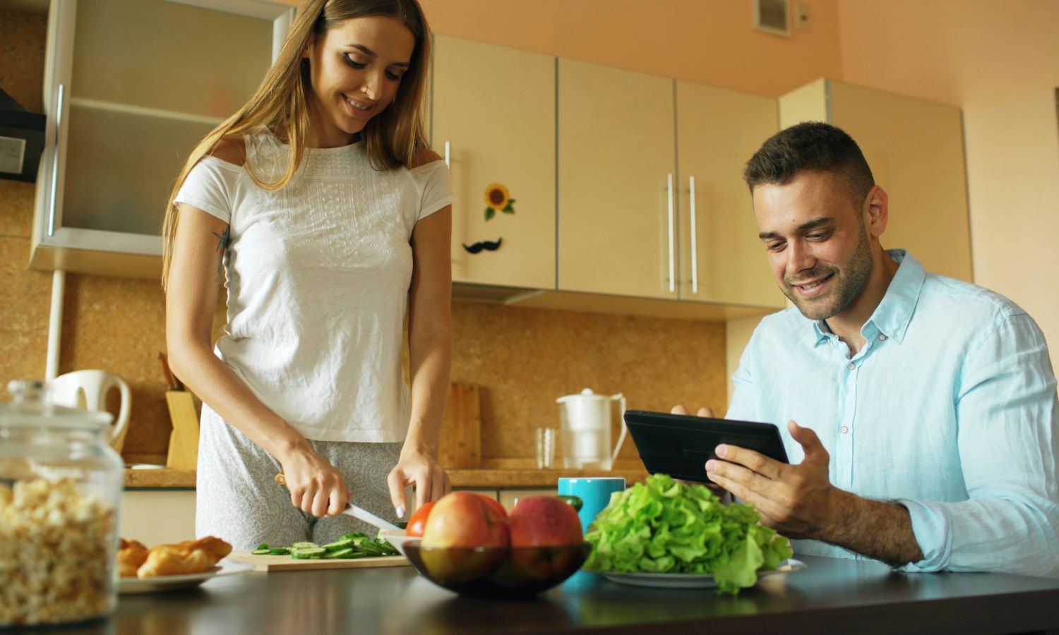 couple in a modern kitchen preparing food together, with the man using a tablet while the woman chops vegetables on the counter