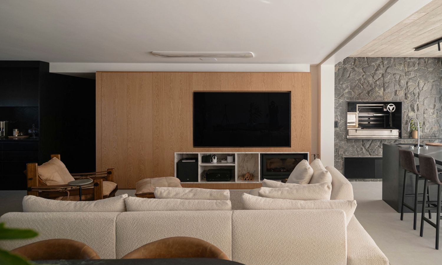 Contemporary living room with a large sectional sofa facing a wall-mounted TV, built-in shelving, warm wood paneling, and an open kitchen area with stone accents.
