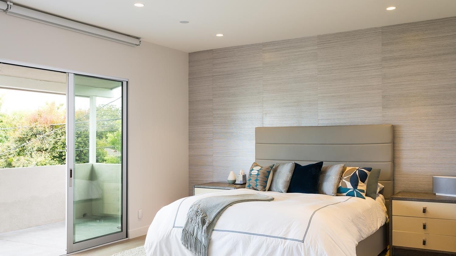 Modern bedroom with a large bed, accent pillows, and a sliding glass door opening to a balcony.